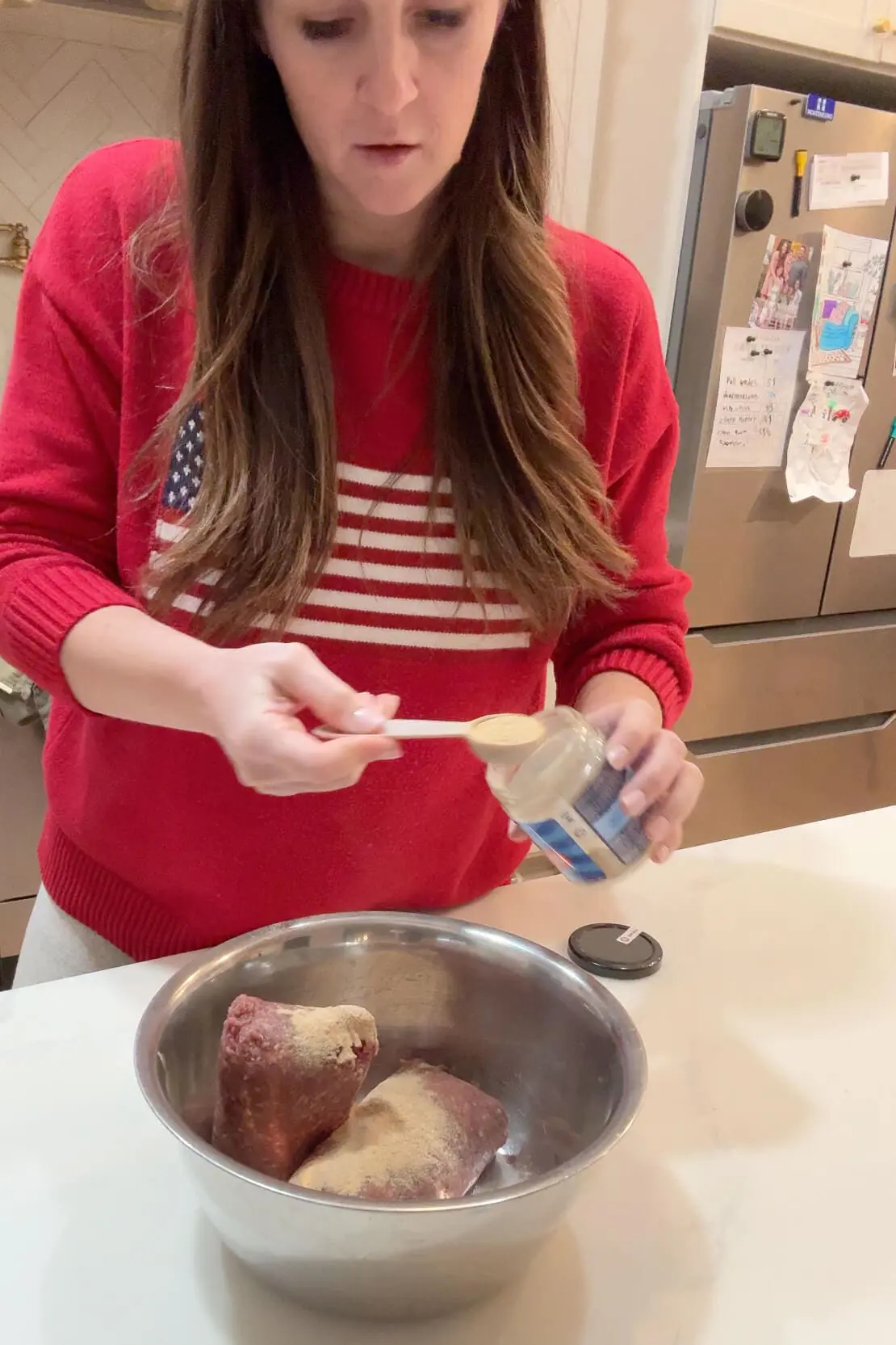 Woman in a red sweater is adding seasoning from a jar into the mixing bowl filled with ground beef. The fridge in the background still showcases family photos and kids’ drawings.